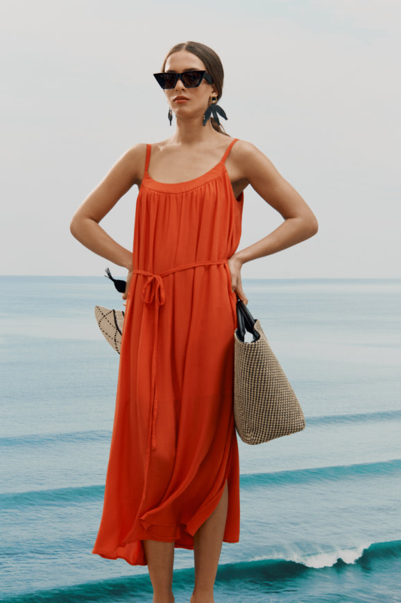 Woman in a flowing dress standing on a beach, holding a woven bag and wearing sunglasses.