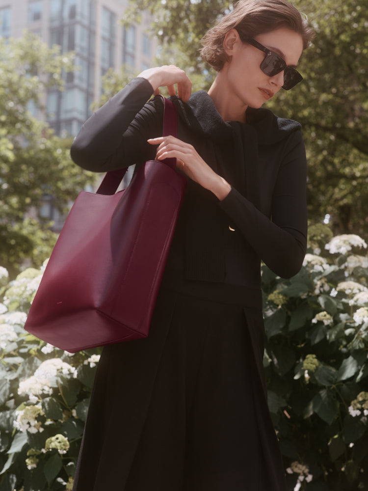 Woman outdoors holding a large tote bag over her shoulder, wearing sunglasses and a long-sleeve outfit.