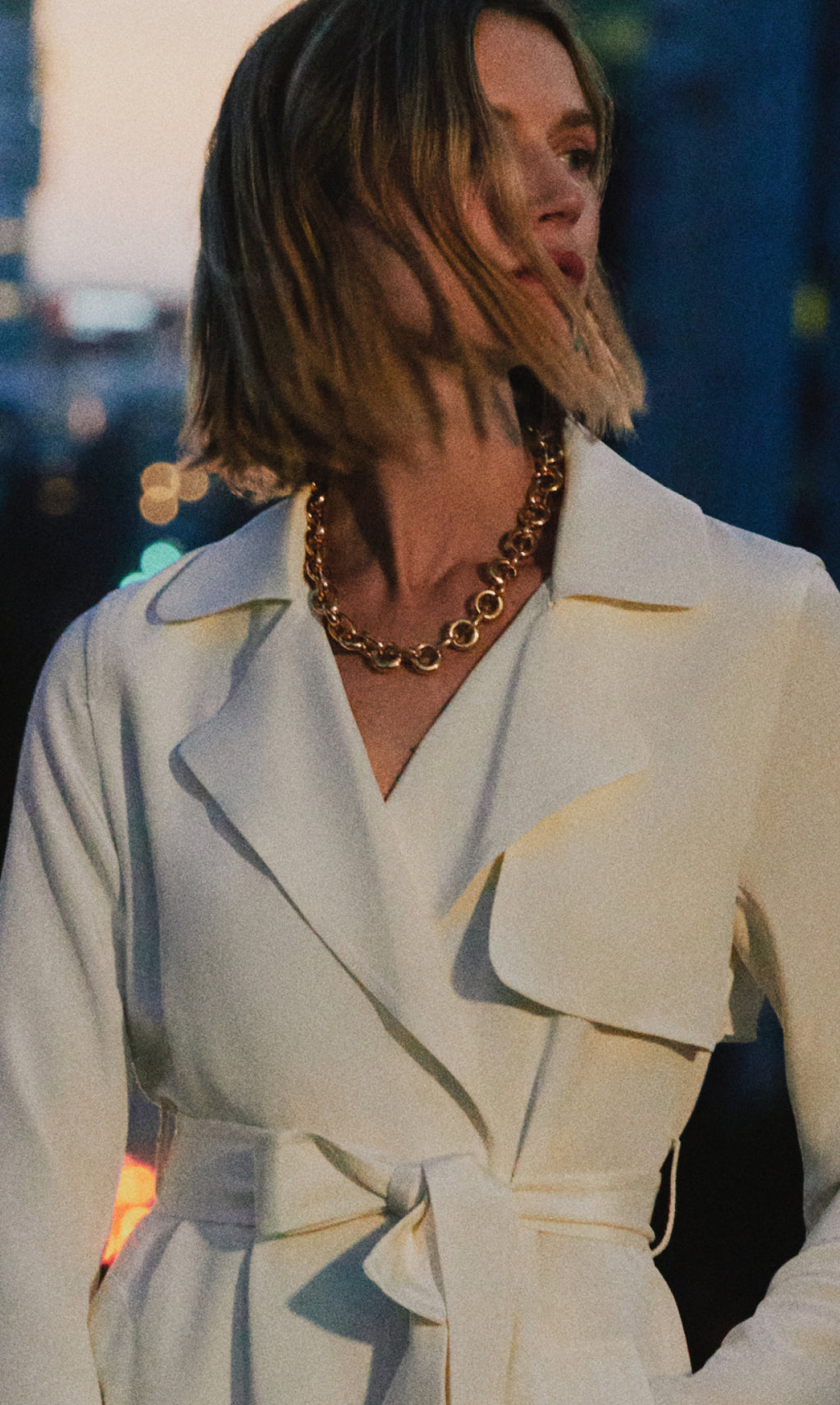 A person wearing a belted trench coat and a chain necklace.