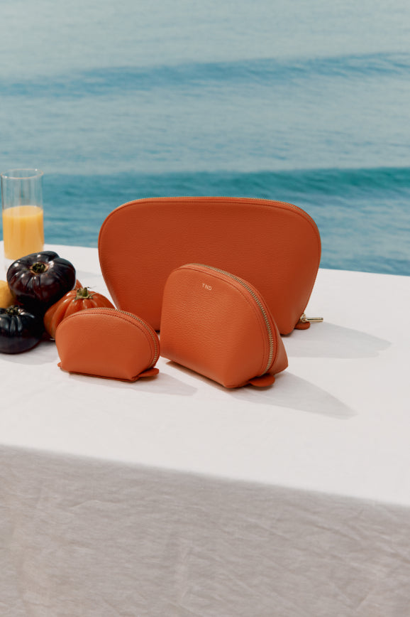 Three cosmetic bags on a table by the sea with fruits and a glass of juice.