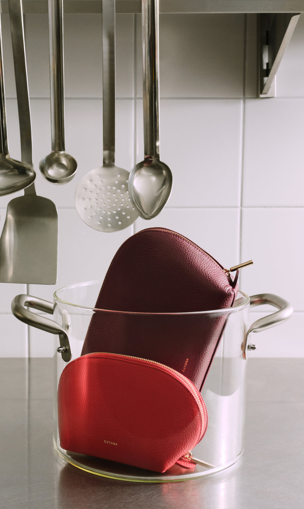 Two purses inside a glass pot on a kitchen counter with hanging utensils.