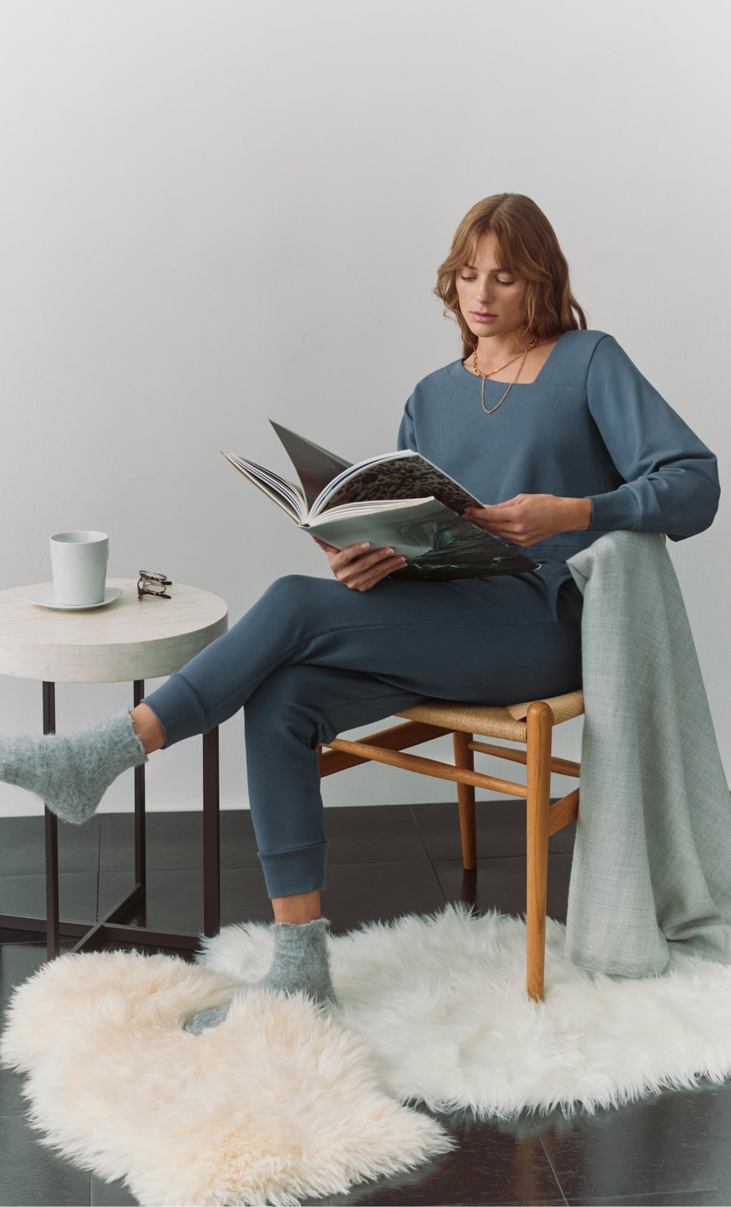 Person reading a book in a chair beside a small round table with a cup and glasses on it.