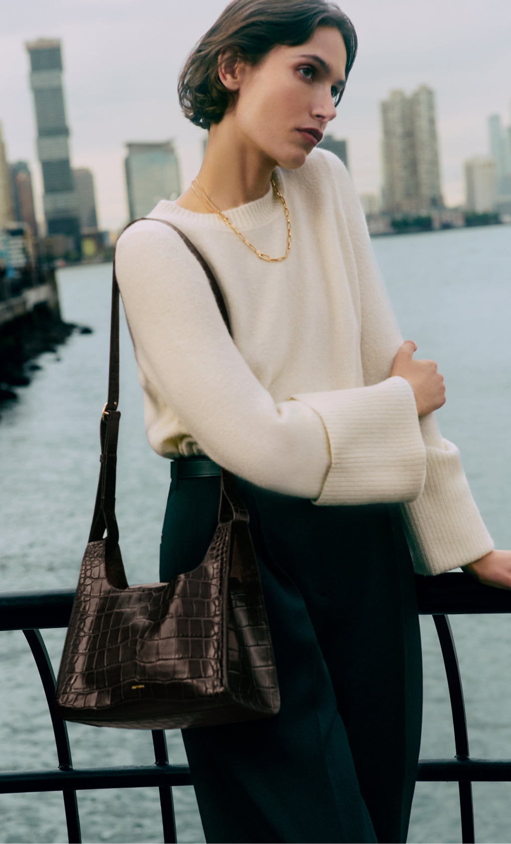 Person leaning on a railing by the water, wearing a sweater and a croc-embossed shoulder bag.