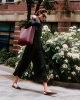 Person walking on a sidewalk with a large bag over shoulder and flower bushes in the background