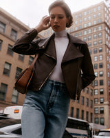 Person in jacket and jeans with a shoulder bag walking in an urban area with tall buildings.