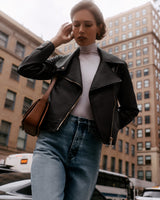 Person wearing a jacket and jeans, holding a bag, walking in an urban area with tall buildings.