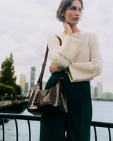 Woman standing by railing holding a croc-embossed leather shoulder bag.