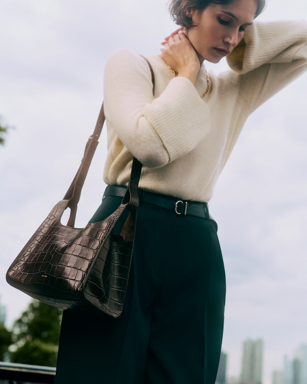 Person with a croc-embossed leather bag over shoulder, wearing a long-sleeved top and high-waisted pants.