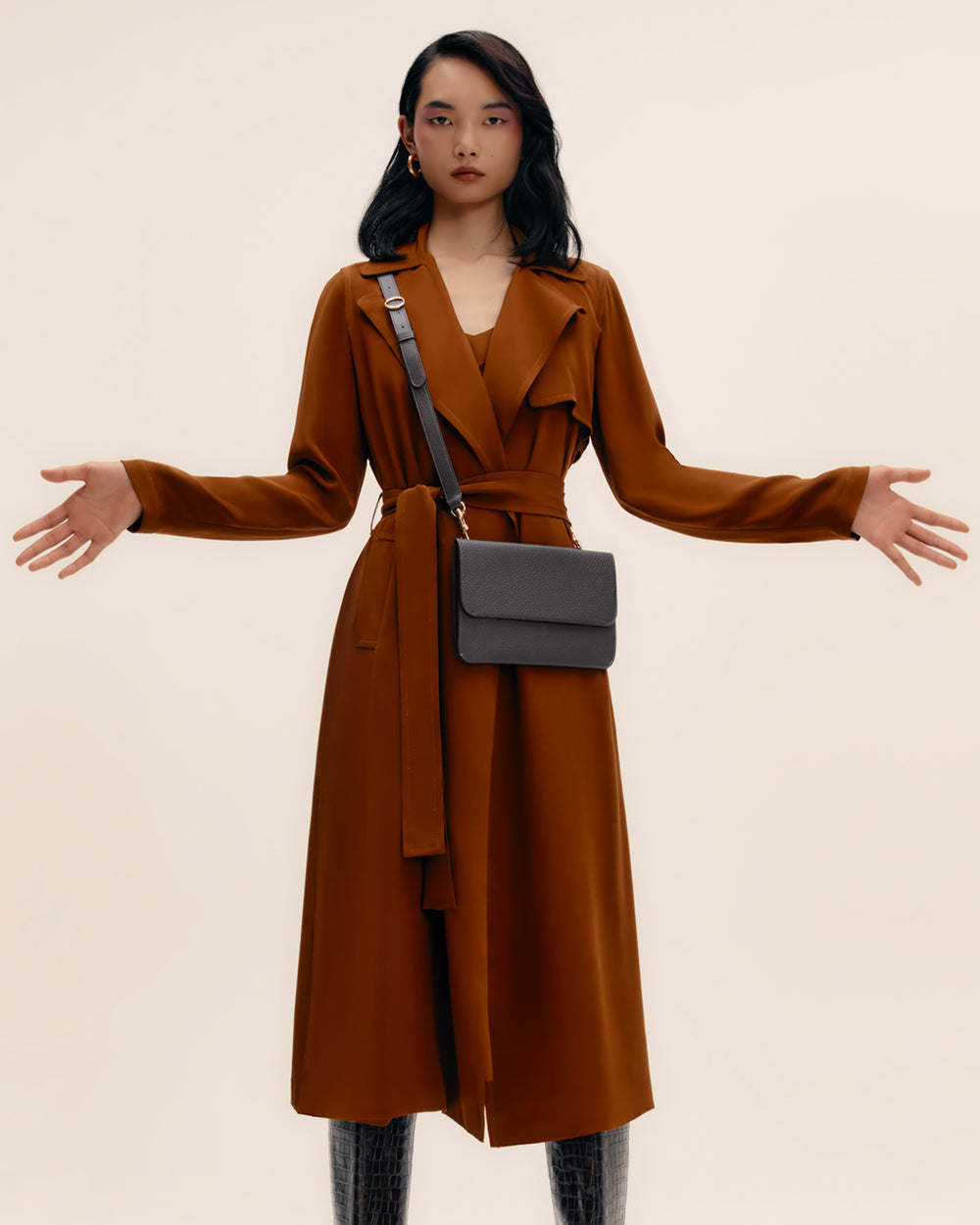 Person wearing a long coat and holding a shoulder bag with arms slightly extended.