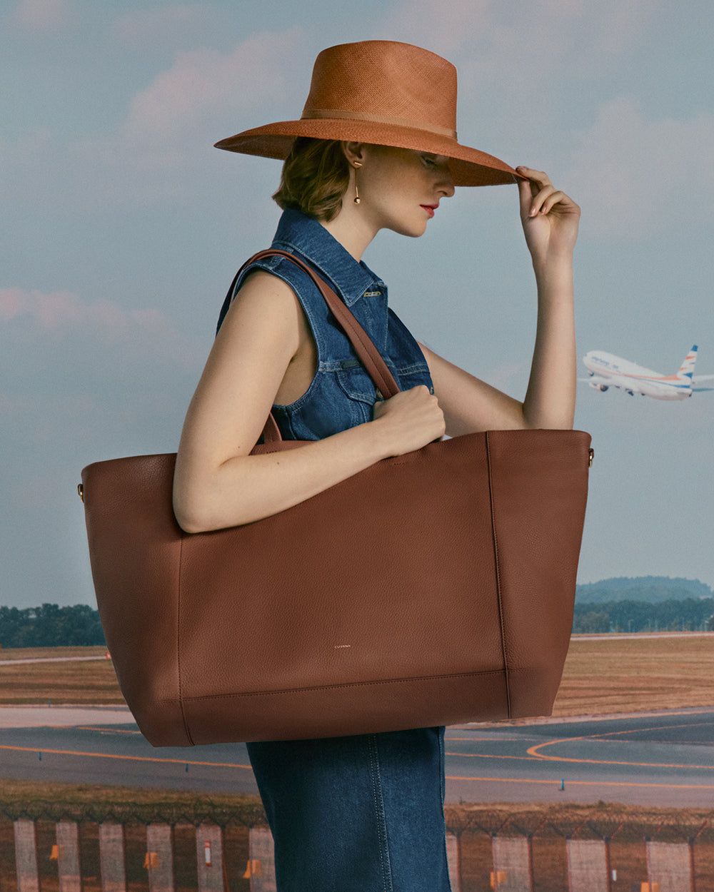 Person in denim dress holding large tote, with an airplane in the background.