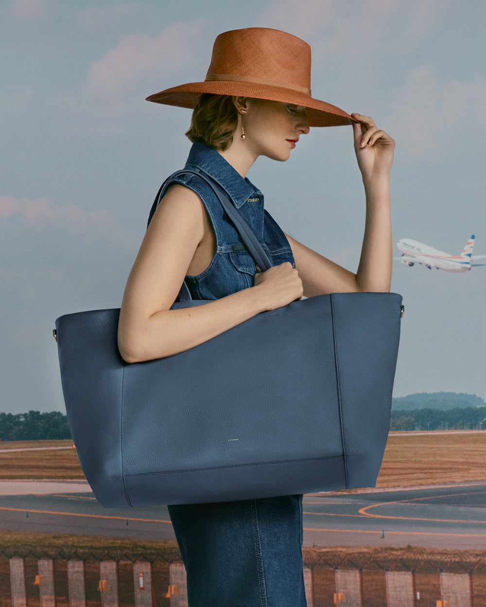 Person in denim dress holding large tote, with an airplane in the background.