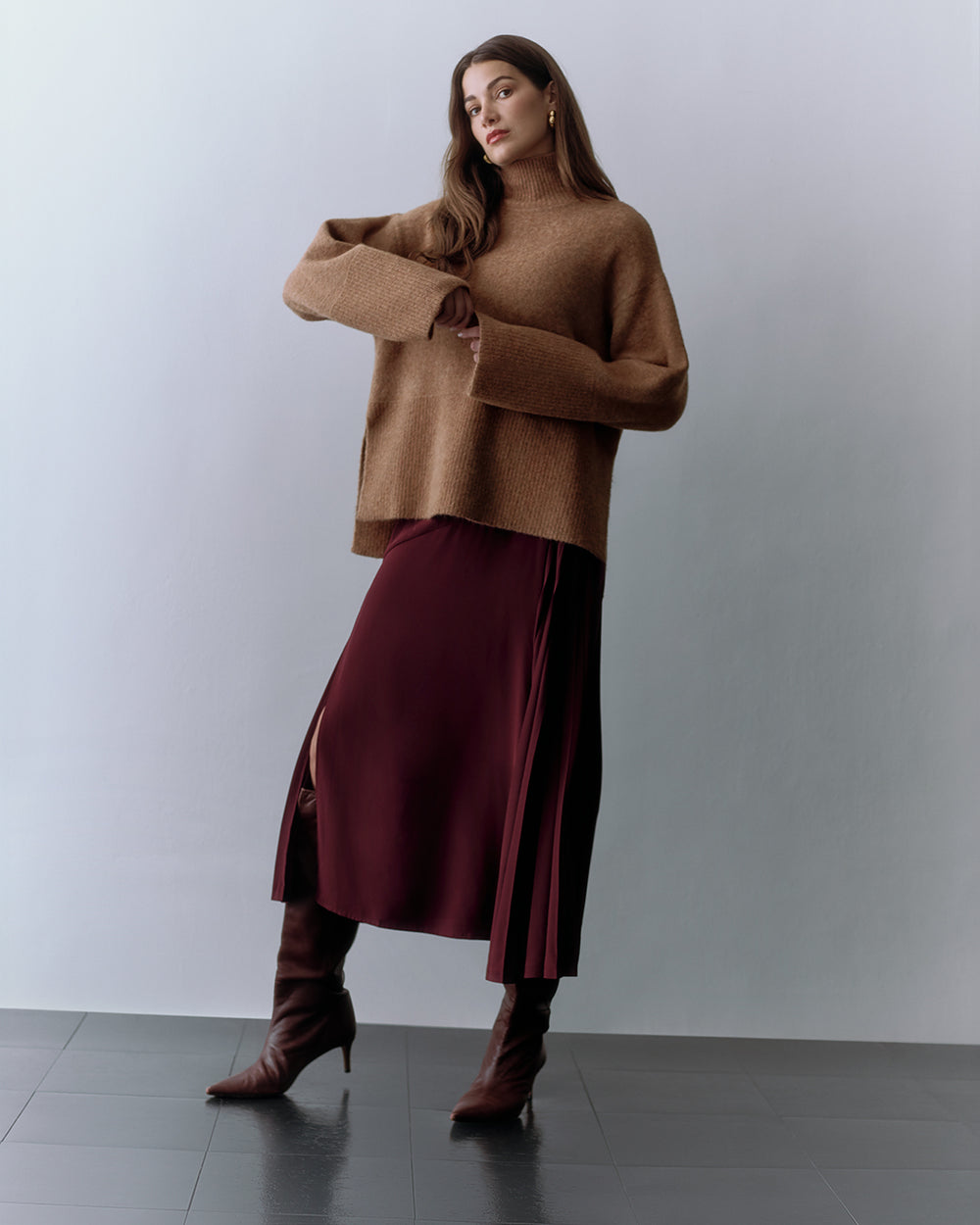 Person wearing a loose sweater, long skirt, and knee-high boots, standing on a tiled floor.