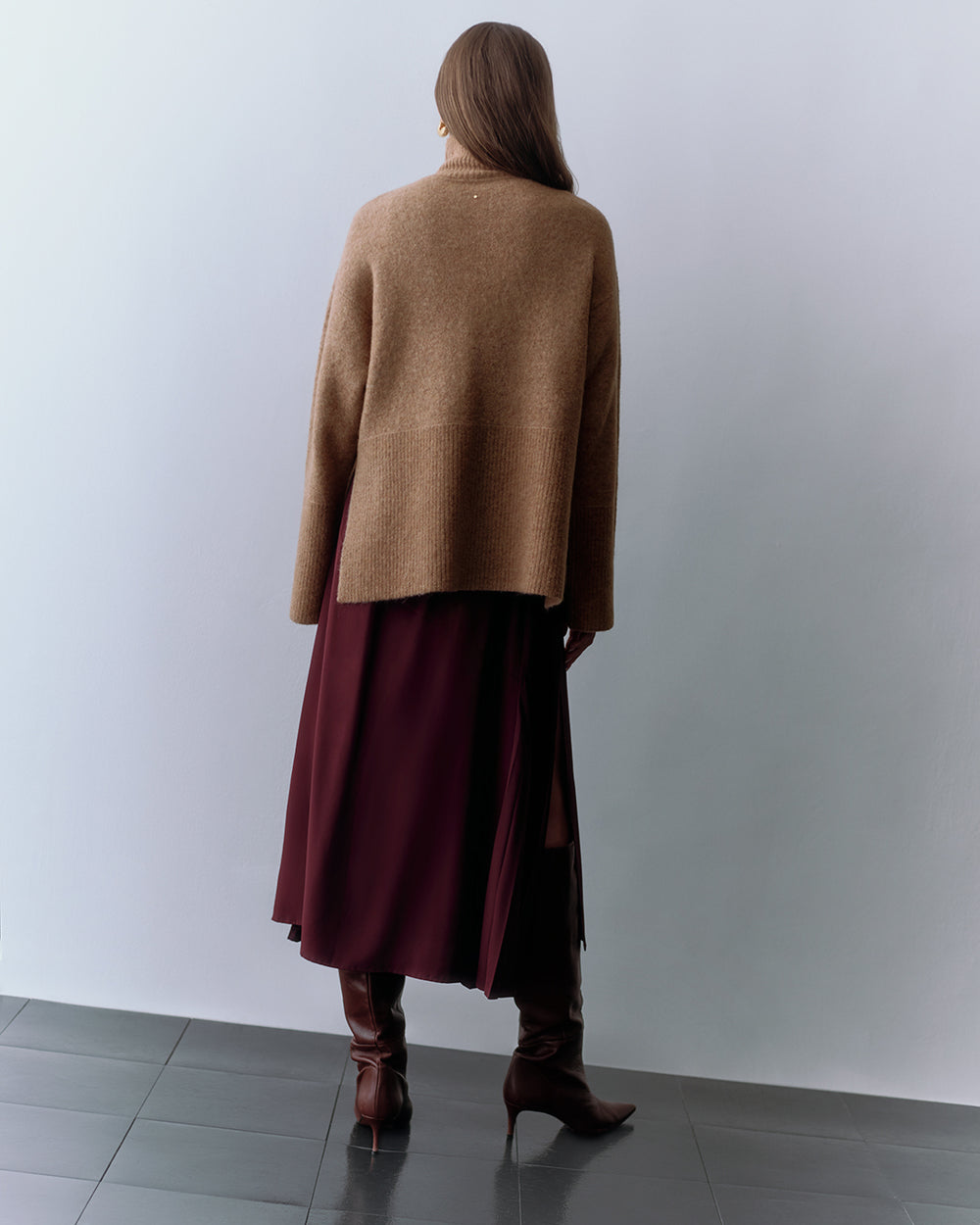 Person wearing a long skirt, loose sweater, and tall boots standing on a tiled floor.