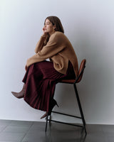 Person sitting on a chair, wearing a sweater and pleated skirt, looking to the side.