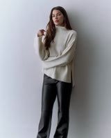 Person wearing a turtleneck sweater and leather pants stands against a plain background.