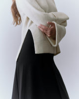 Person standing sideways wearing a long-sleeve top and skirt, arms crossed.