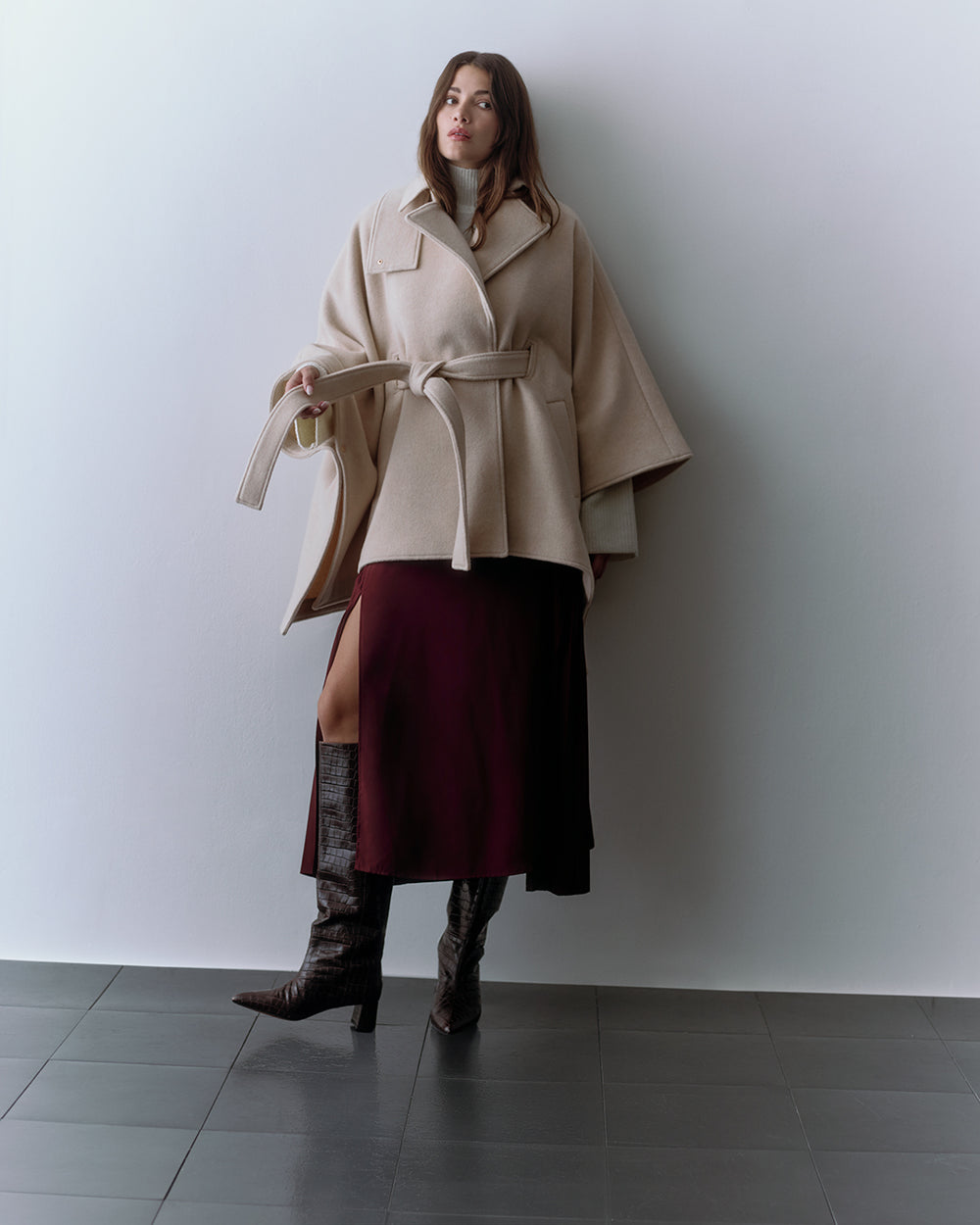 A model in a belted cape and skirt with knee-high boots stands against a wall.