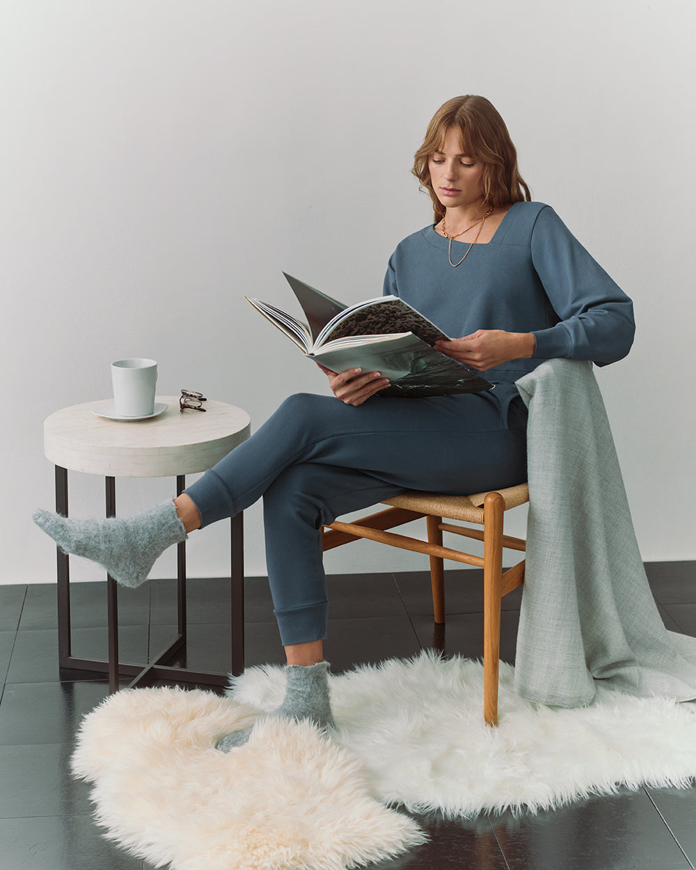 Person wearing matching sweater with an asymmetrical neckline and joggers, sits and reads with a blanket and a mug on a side table.  