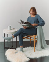 Person wearing matching sweater with an asymmetrical neckline and joggers, sits and reads with a blanket and a mug on a side table.  