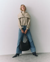 Person in a cape and jeans holding a handbag standing against a plain background.