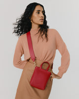 Woman with long curly hair standing with hand on hip, wearing a turtleneck and holding a small bag.