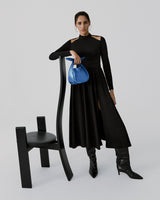 Woman standing next to a chair, holding a handbag, wearing a long dress and boots.