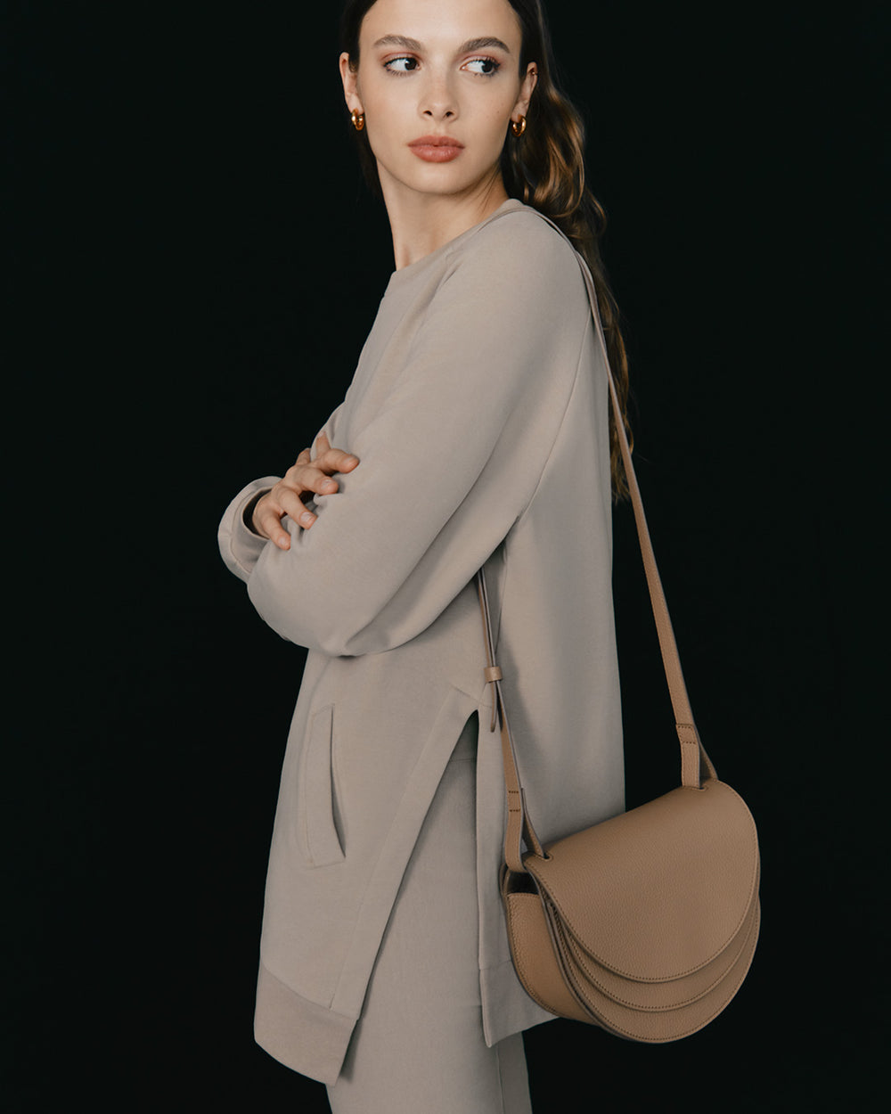 Person wearing a long sleeve top and semicircular bag standing sideways with arms crossed