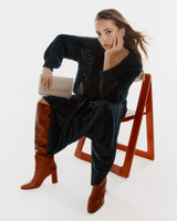 Woman sitting on a chair, posing with one hand on her face and holding a clutch.