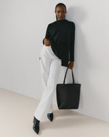 Person standing against a wall wearing a top, pants, and holding a tote bag.