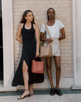 Two women standing by a wall, one holding a bag, both looking to the side.