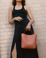 Woman with a handbag standing against a brick wall, holding sunglasses.