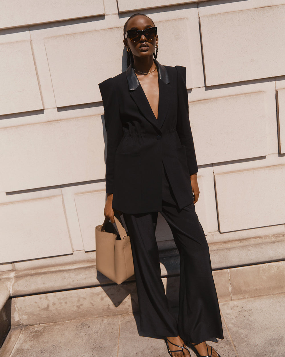 Woman in a suit standing against a wall with a handbag and sunglasses.