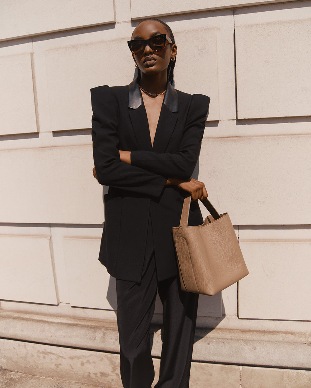 Woman in a suit holding a bag, leaning against a wall with sunglasses on.