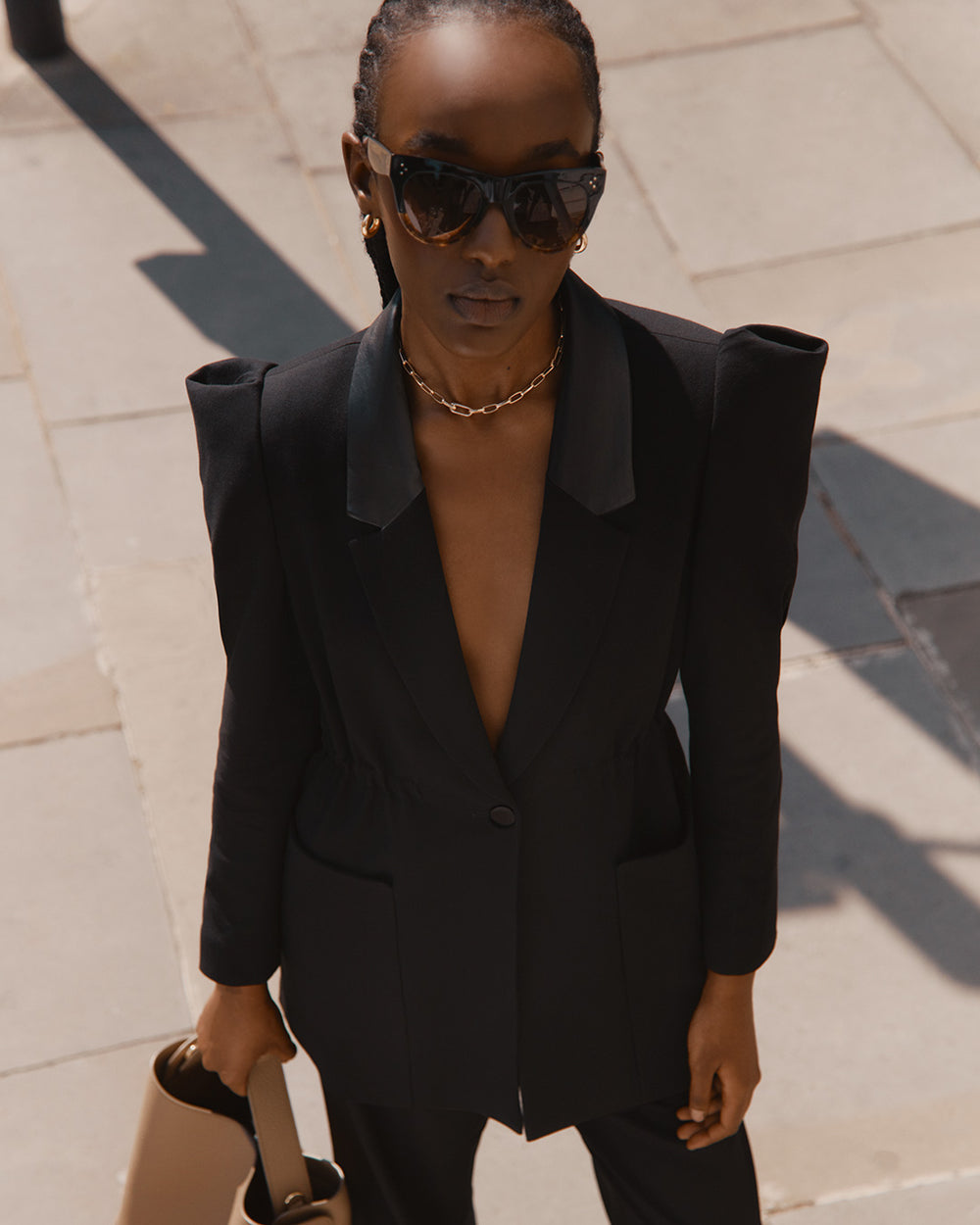 Woman in a suit standing outdoors with sunglasses and a handbag.