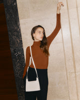 Young woman standing indoors, arm raised, carrying a shoulder bag.