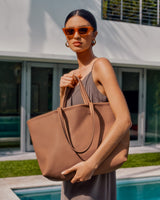 Woman with sunglasses holding a large tote bag standing by a pool.