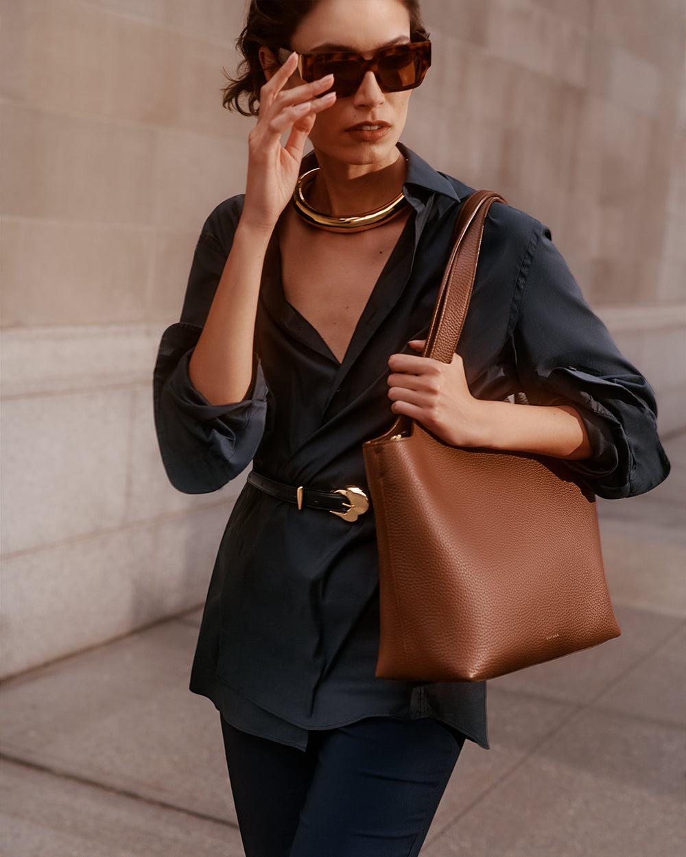 Person wearing sunglasses, a collared top, pants, and a chunky necklace, holding a shoulder bag