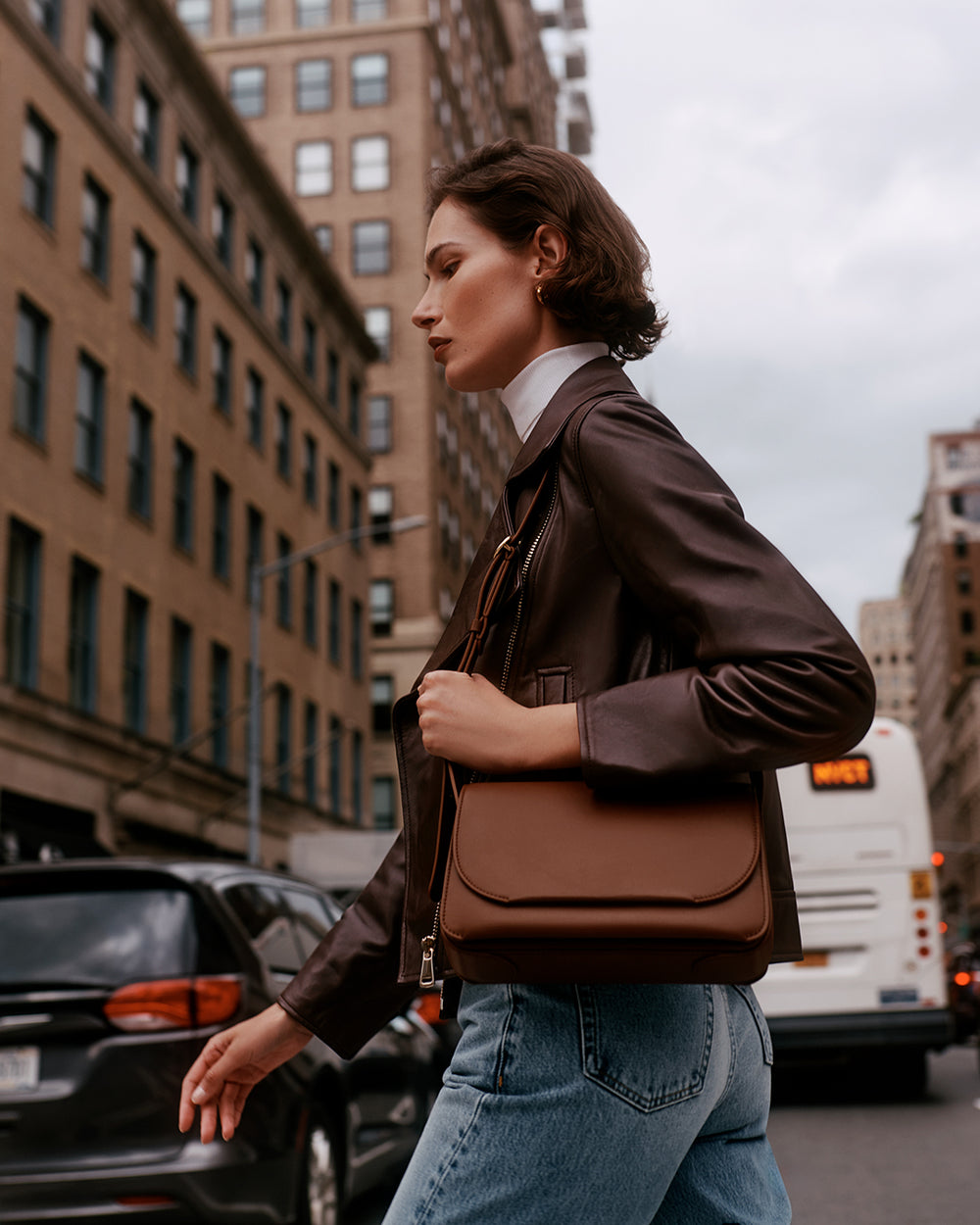 Person walking down a city street carrying a handbag, with buildings and cars in the background