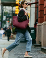 Person walking in the city holding a large handbag, wearing sunglasses, a coat, and jeans.