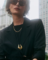 Person wearing a suit and sunglasses, with a necklace, in an urban setting.