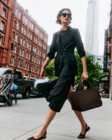 Person walking on city sidewalk carrying large bag, wearing sunglasses and jacket.