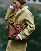 Person wearing a trench coat holding a large tote bag standing on a city street.