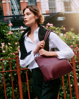 Woman standing near fence holding a shoulder bag, with a sweater draped over her shoulders