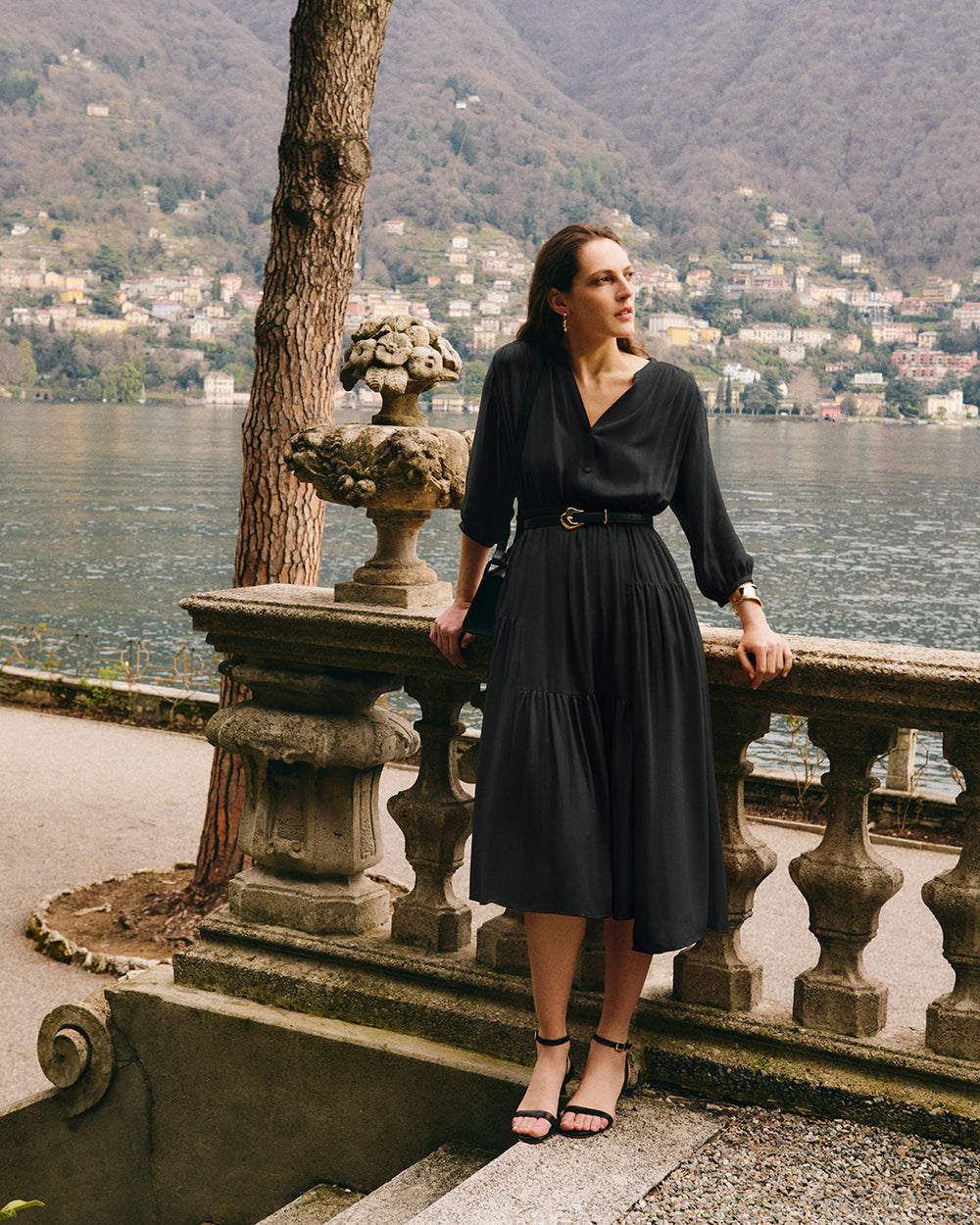 Person in dress stands by stone railing with lake and hills in background.