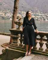 Person in dress stands by stone railing with lake and hills in background.