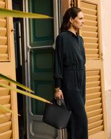 Person in jumpsuit holds bag, standing near window shutters.