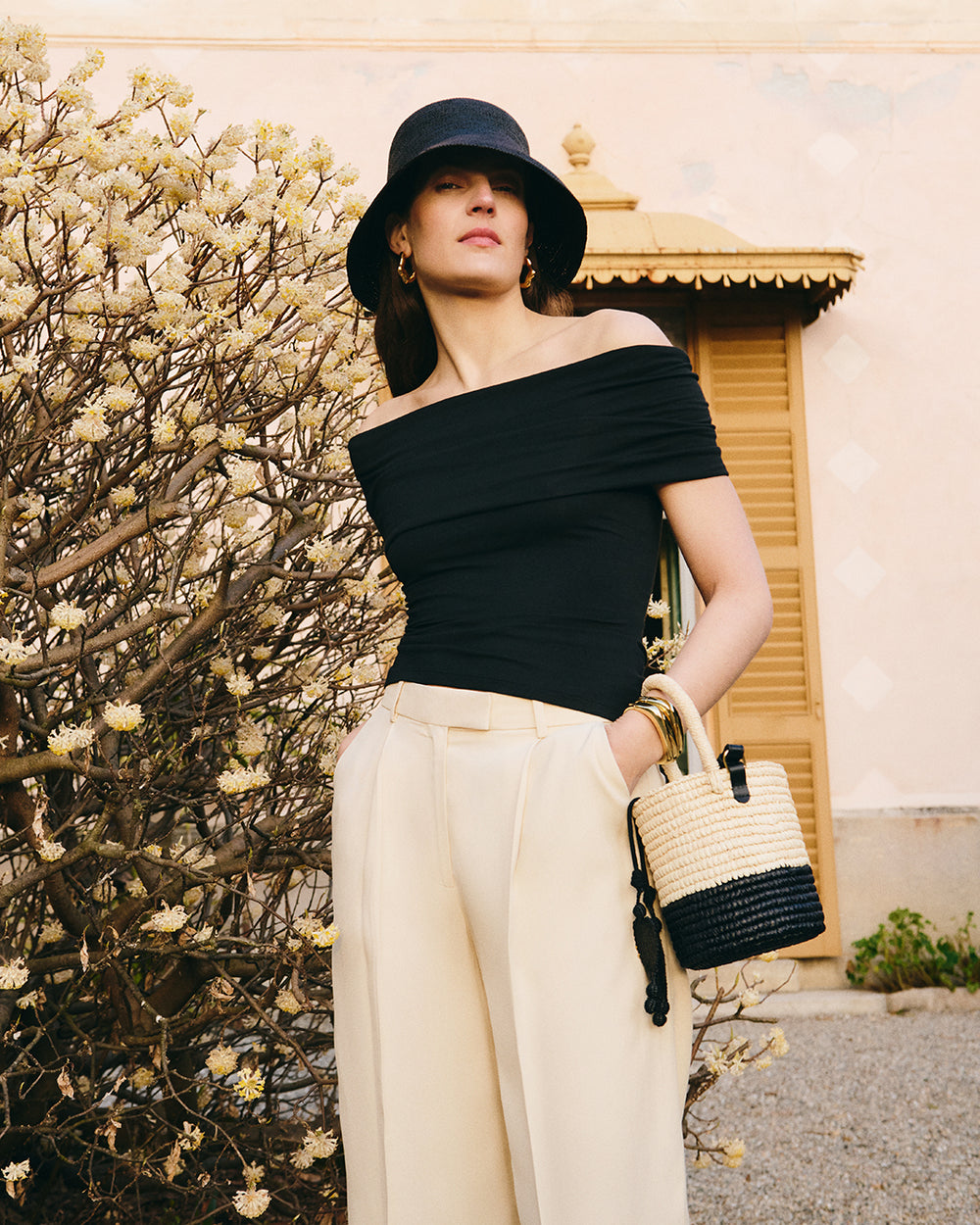 Person wearing off-shoulder top and wide-leg trousers, holding a bag and standing near a bush.