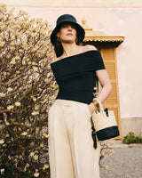 Person wearing off-shoulder top and wide-leg trousers, holding a bag and standing near a bush.
