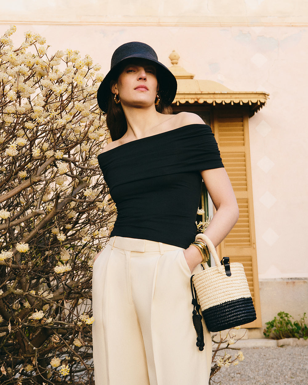 Woman with hat and shoulder bag poses by a bush and building with shutters.