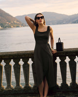 Person in a dress leaning on a stone railing by a lake with hills in the background